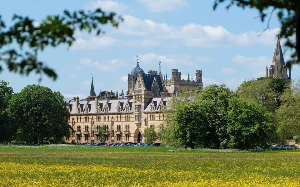 A historic Oxford university college building overlooking a sunlit meadow